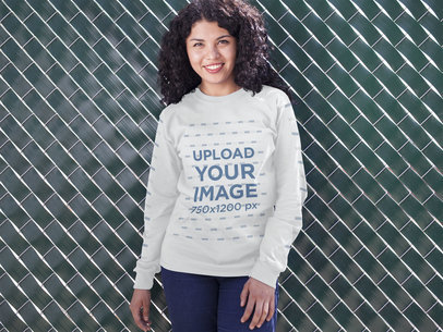 Mockup of a Young Woman Wearing a Crewneck Sweatshirt while Standing Against a Green Fence Outdoors
