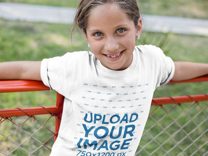 Smiling Little Girl Wearing a T-Shirt Mockup While Lying Against a Red Railing a16170