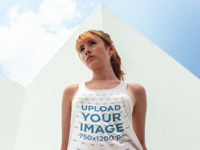 Mockup of a Beautiful White Girl Wearing a Bella Canvas Tank Top While Near a White Building