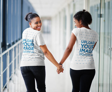 Back-View Tee Mockup of Two Smiling Women Holding Hands in a Hallway