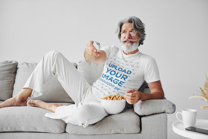 T-Shirt Mockup of a Bearded Senior Man Sitting on His Couch with a Bowl of Snacks