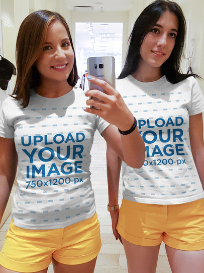 Two Women Taking a Selfie Against a Mirror Wearing Round Neck Tees Mockup at a Fitting Room