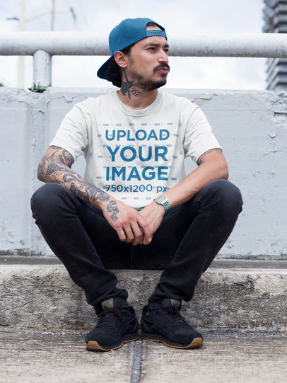 Mockup of a Tattooed Hispanic Man Wearing a Round Neck Tee While Sitting Down on a Sidewalk