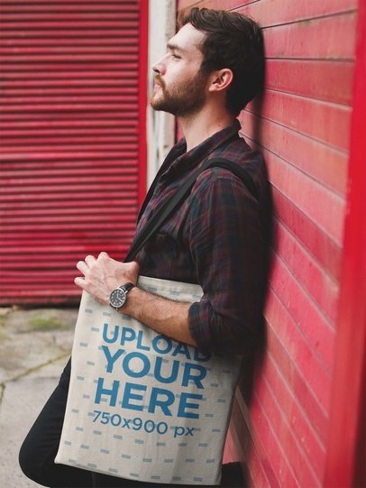 Man Holding a Tote Bag Mockup While Lying Against a Red Wall