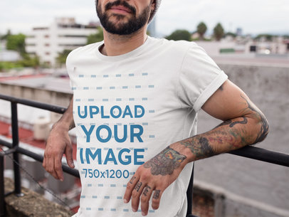 Cropped Face Tattooed Man Wearing a T-Shirt Mockup While Lying Against a Fence on a Rooftop a17027