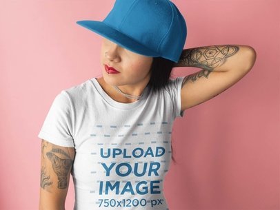 Tattooed Woman Wearing a T-Shirt Mockup and a Hat Against a Pink Background