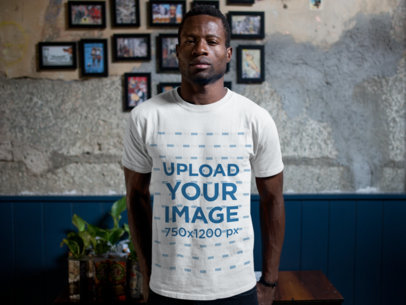 Tshirt Mockup of a Man Standing Against a Wall with Framed Pictures