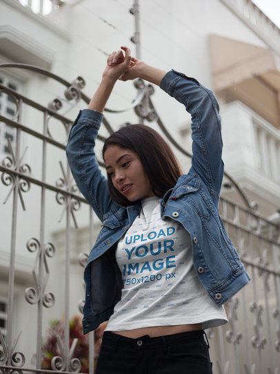 Asian Woman Wearing a Round Neck Tee Mockup and a Denim Jacket While Lifting her Arms Outdoors