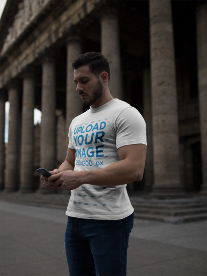 Strong Man Wearing a T-Shirt Mockup While Using his Phone Near a Historic Building a17662
