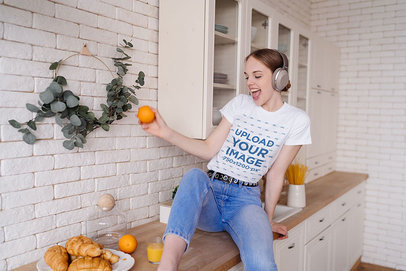 T-Shirt Mockup Featuring a Joyful Young Woman Holding an Orange in the Kitchen