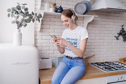 T-Shirt Mockup Featuring a Woman With Headphones Sitting on a Kitchen Counter