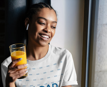T-Shirt Mockup Featuring a Woman Drinking Orange Juice by a Window