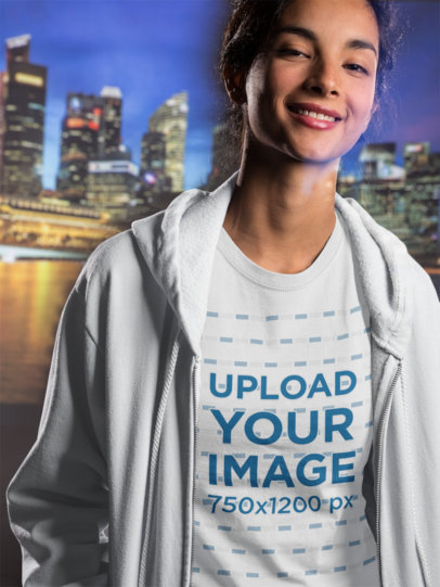 Smiling Woman Wearing a T-Shirt Mockup and a Hoodie Against a City Landscape