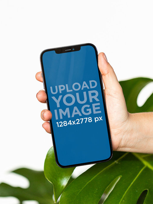 Mockup of a Hand Holding an iPhone 12 Pro Max in Front of a Plant