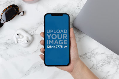 Mockup of a Hand Holding an iPhone 12 Pro Max Over a Marble Desk