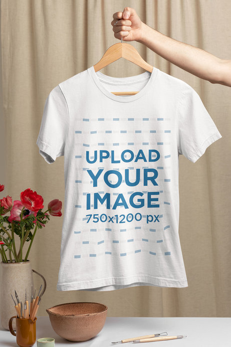Mockup of a Woman's Hand Holding a Hanger with a T-Shirt Over a Table with Flowers