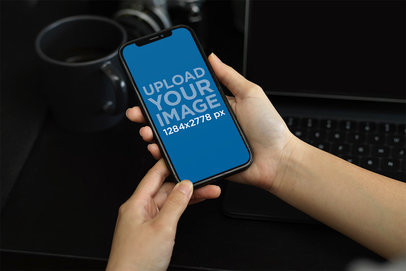 Mockup of a Woman Holding an iPhone 13 Featuring a Black Desk in the Background
