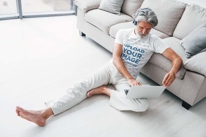T-Shirt Mockup of an Elderly Man Sitting in His Living Room While Using a Laptop