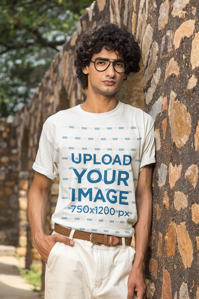 T-Shirt Mockup of a Man with Contact Glasses Leaning on a Stone Wall