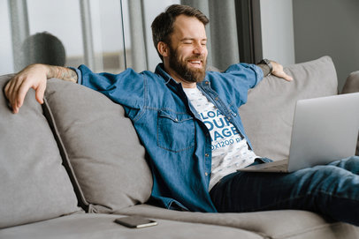 Tee Mockup of a Bearded Man Sitting on a Couch with a Computer on His Lap