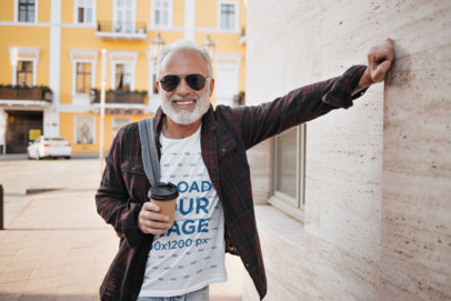 T-Shirt Mockup of a Senior Man Leaning Against a Wall and Holding a Cup of Coffee