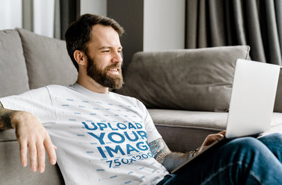 Round-Neck Tee Mockup of a Man with an Arm Tattoo Using a Laptop by a Couch