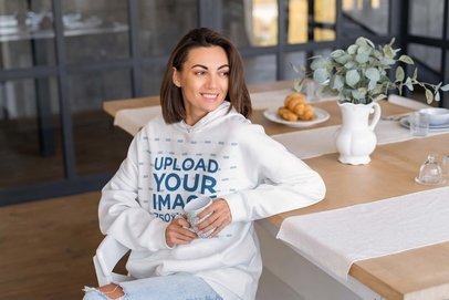 Hoodie Mockup of a Happy Woman Sitting on the Dining Room Table