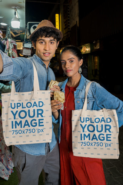 Tote Bag Mockup of a Couple Taking a Selfie while Eating Street Food