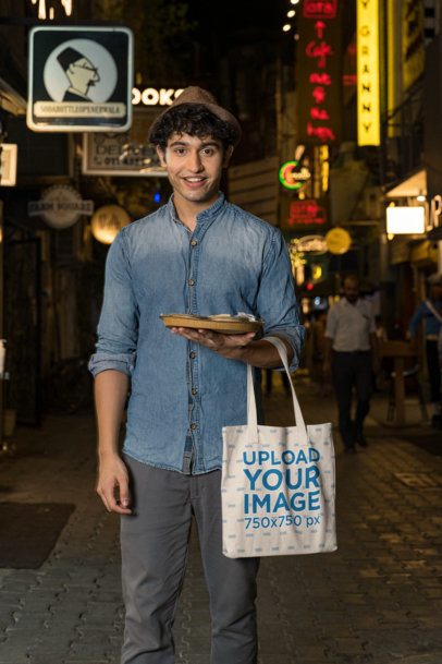 Tote Bag Mockup Featuring a Man on a Street Holding a Dish of Food 