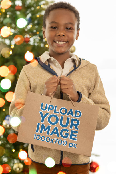 Mockup of a Smiling Kid Holding a Gift Bag in Front of a Christmas Tree