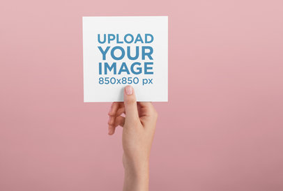 Postcard Mockup of a Female Hand Holding a Square Postcard Over a Wall