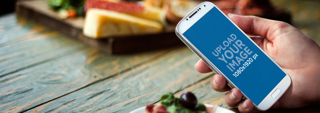 Mockup of a Person Holding a Samsung Galaxy Featuring Food and a Wooden Table