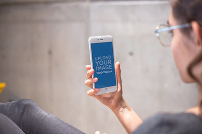 Mockup of a Girl with Glasses Holding a Silver iPhone 8 Plus Near a Concrete Wall a21284
