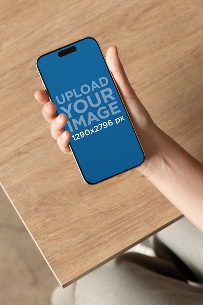 Mockup of a Woman Holding an iPhone 15 Pro Max Against a Wooden Surface