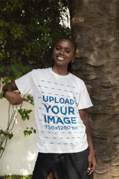Mockup of a Smiling Woman Pointing at Her T-Shirt While Posing Against a Tree