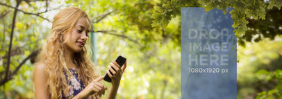 Beautiful Mockup of Blonde Woman Using Her Smartphone Below a Tree