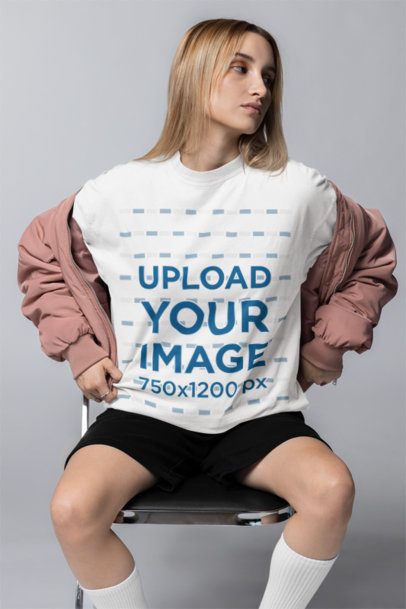 Oversized T-Shirt Mockup Featuring a Woman Wearing a Jacket and Sitting on a Chair in a Studio