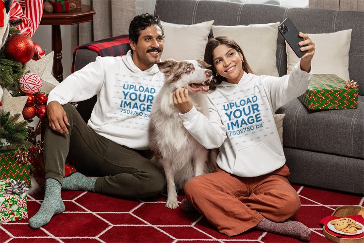 Christmas Mockup Featuring a Happy Couple in Gildan Hoodies Taking a Selfie With a Dog in a Living Room