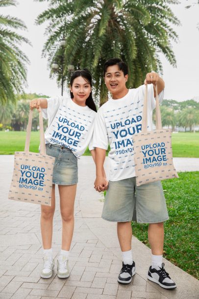 Oversized T-Shirt Mockup Featuring a Cheerful Couple Showing Their Tote Bags in the Park