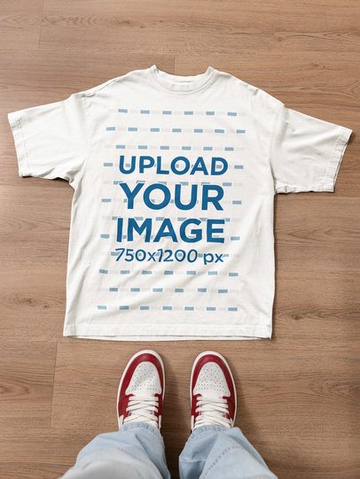 Mockup of an Oversized T-Shirt Lying on a Wooden Floor Featuring a Man's Sneakers