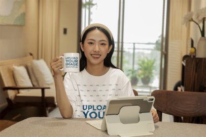 Oversized T-Shirt Mockup Featuring a Happy Woman Holding a Coffee Mug While Using a Tablet