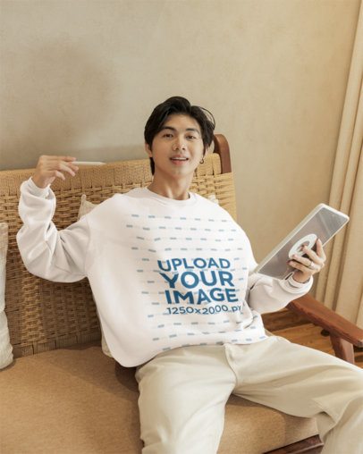 Sweatshirt Mockup Featuring a Smiling Man Sitting in a Cozy Chair With a Tablet