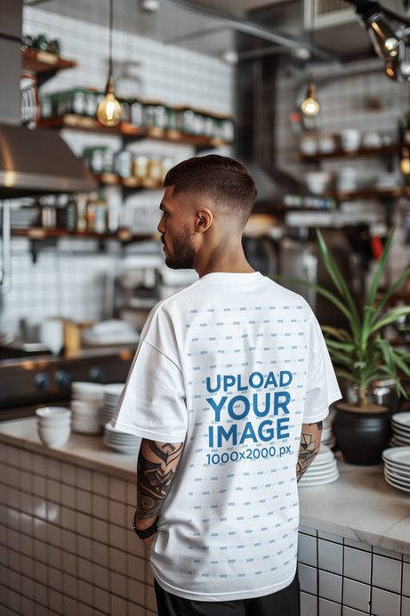 Back-View T-Shirt Mockup Featuring an AI-Created Tattooed Man in a Restaurant