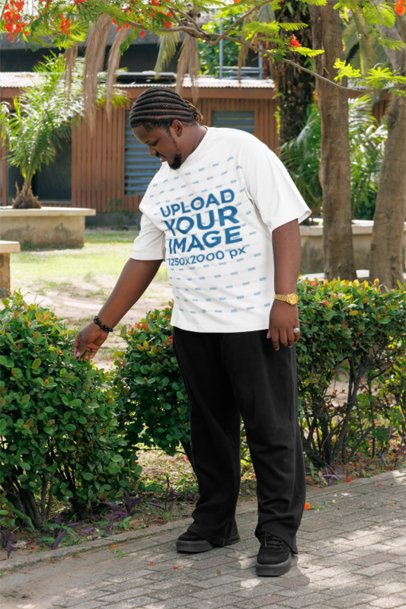 Oversized T-Shirt Mockup of a Smiling Man Walking and Touching Some Plants in a Park