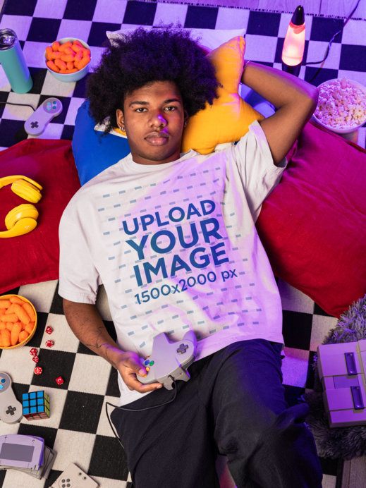 Oversized T-Shirt Mockup Featuring a Serious Man Relaxing on the Floor With Gaming Stuff