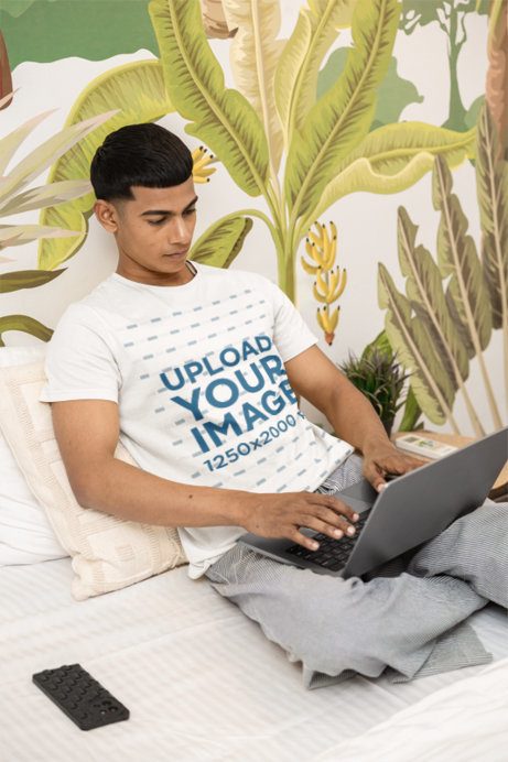 T-Shirt Mockup of a Man Resting on a Cozy Bed While Checking His Laptop