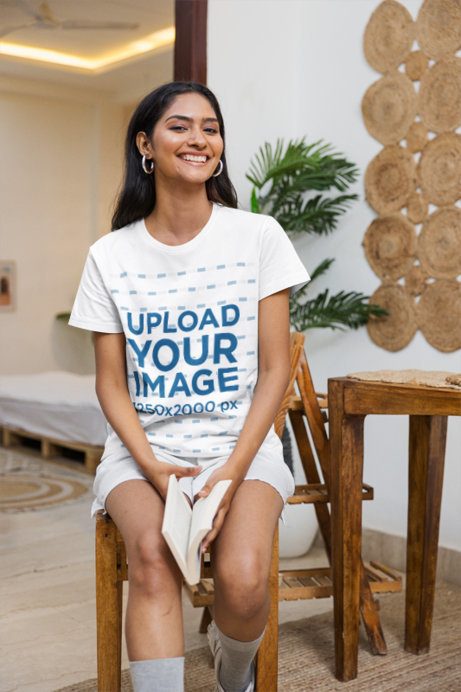 T-Shirt Mockup featuring a Smiling Young Woman holding an Open Book on a Wooden Stool