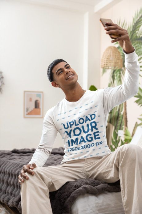 Long Sleeve T-Shirt Mockup featuring a Young Man Taking a Selfie While Sitting on a Bed