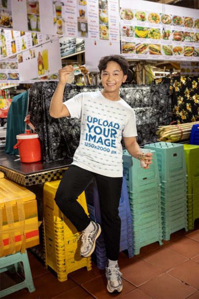 T-Shirt Mockup Featuring a Smiling Young Man Eating Food at a Street Market Stall