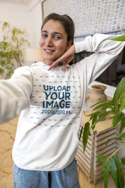 Hoodie Mockup Featuring a Smiling Young Woman Taking a Selfie in a Bright Room With Plants